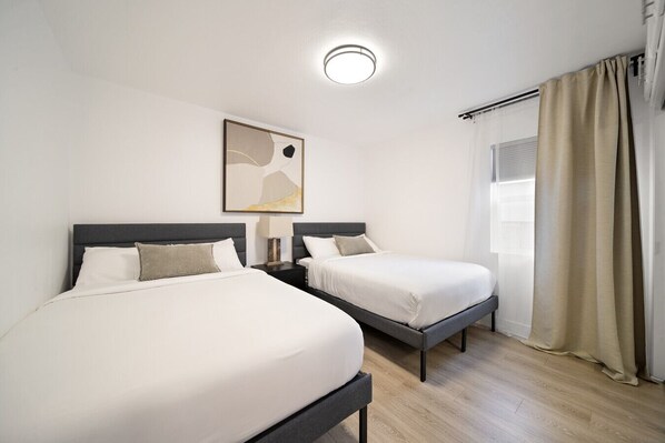 A hotel room with two beds, a ceiling light, a window with curtains, and a framed artwork on the wall.