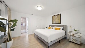 A modern bedroom with a large bed, a bedside table, a wall-mounted artwork, and a potted plant.