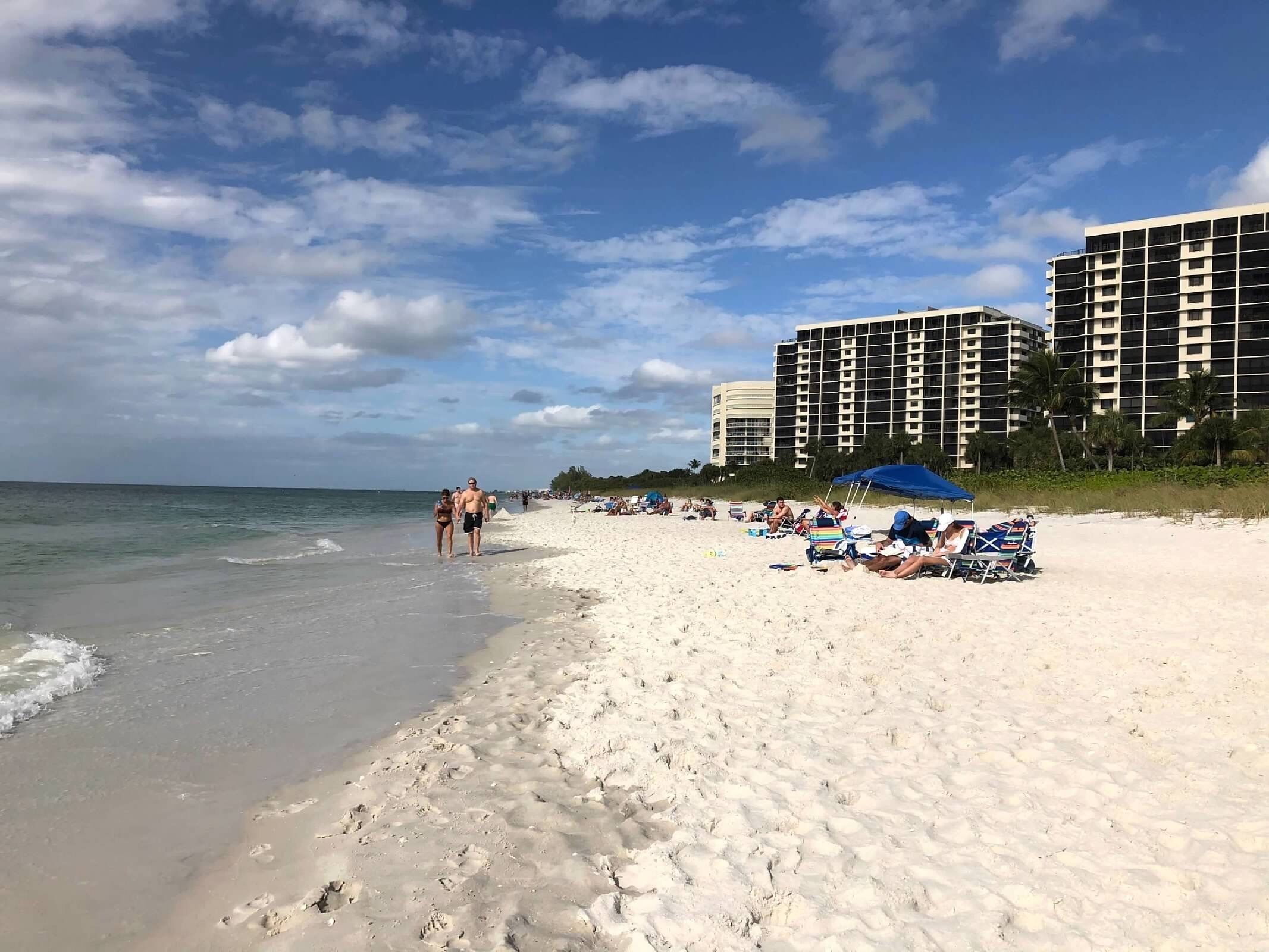 Een strand met wit zand, een heldere lucht en enkele mensen die wandelen. Achtergrondgebouwen en een paar strandparasols.