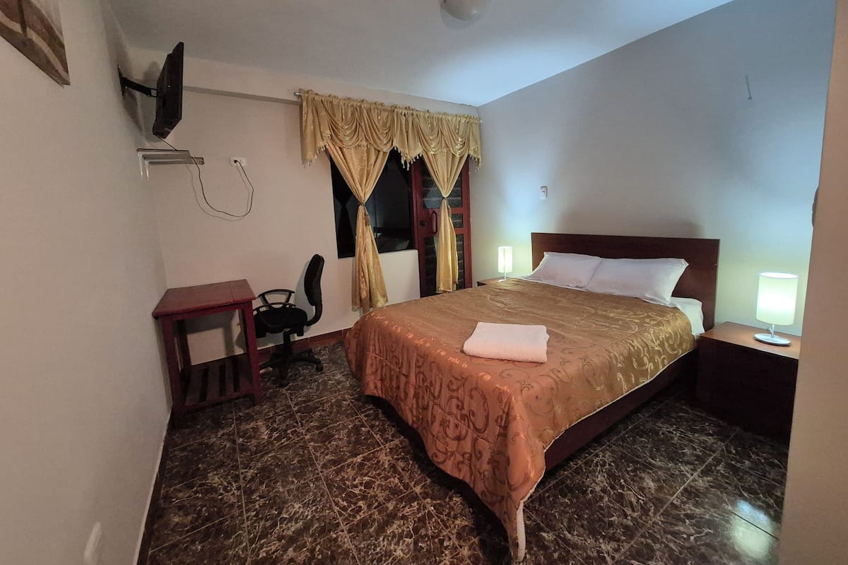 A hotel room with a bed, a desk, a chair, a TV mounted on the wall, and a window with curtains.