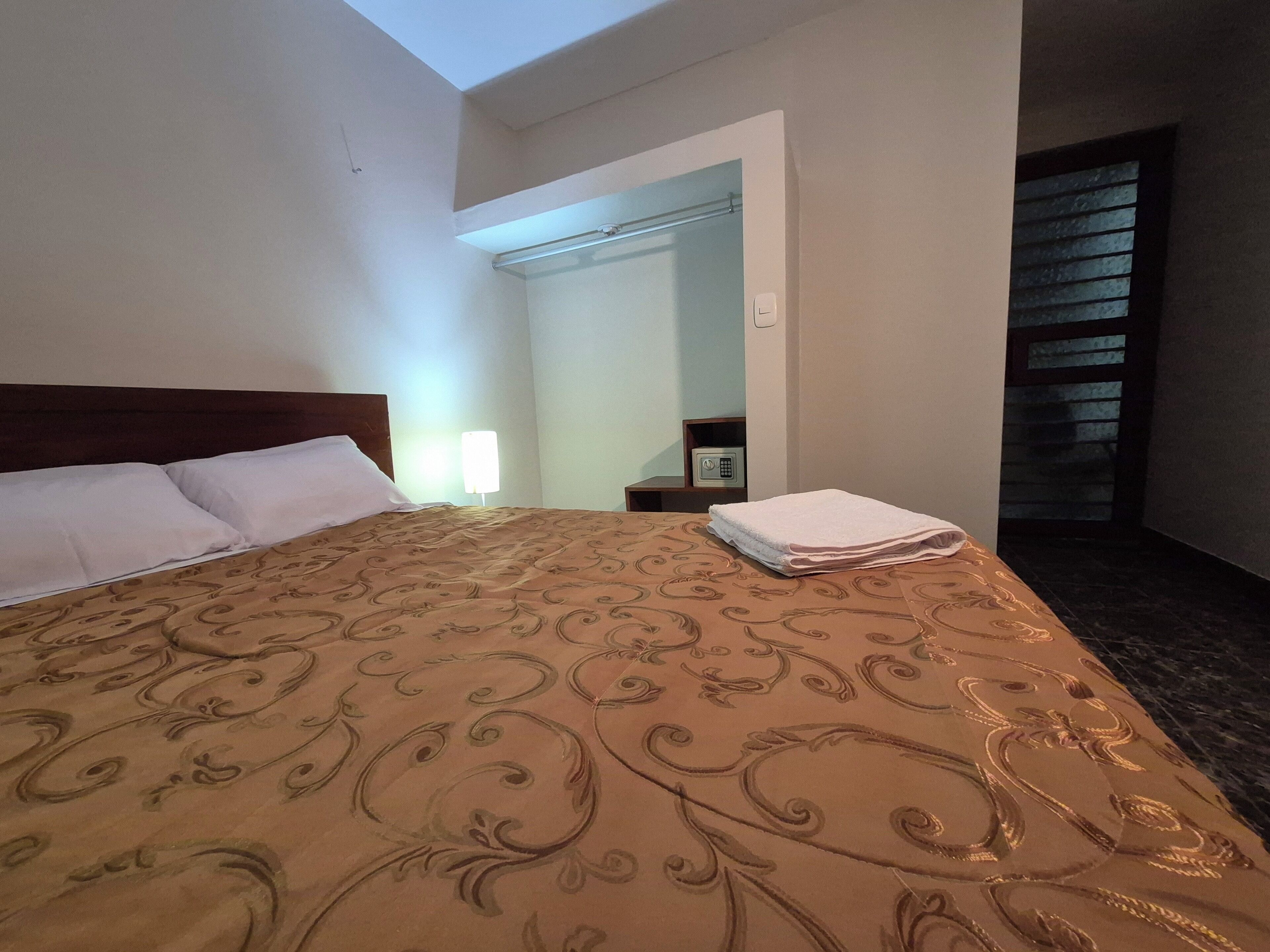 Habitación de hotel con una cama grande, una mesita de noche, una lámpara y vistas al exterior a través de una ventana.