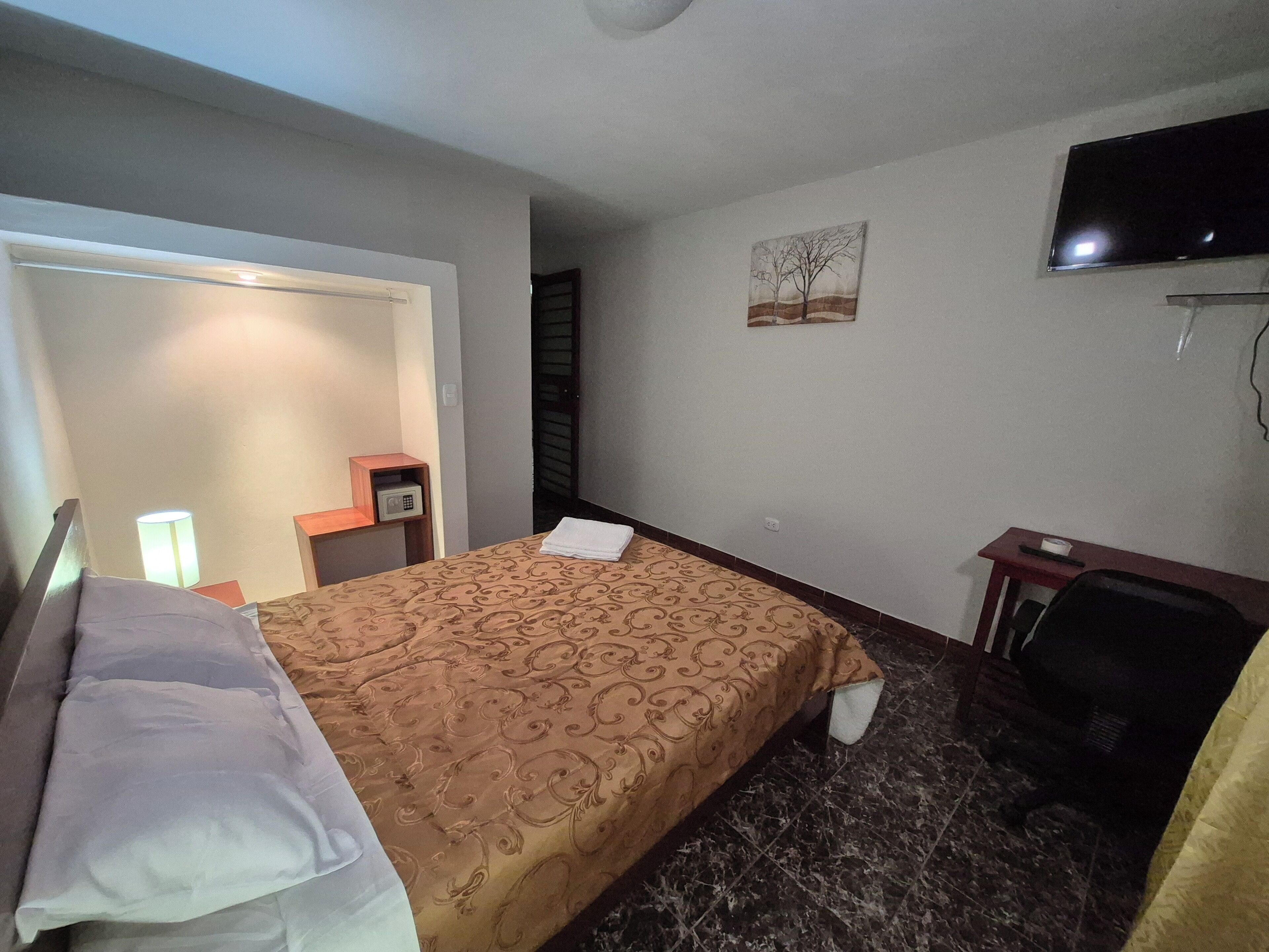 A hotel room with a bed, a desk, a chair, a television, and a painting on the wall.