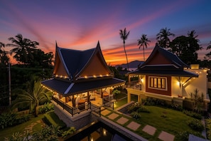 A two-story house with a balcony and a covered patio area, surrounded by a garden and a body of water.