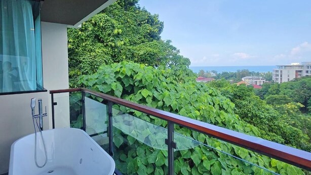 A balcony with a glass railing, a white bathtub, and a view of lush greenery and distant buildings. - Brand New 2 Bed Seaview Apartment Utopia Karon (karon)
