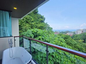 A balcony with a glass railing, a white bathtub, and a view of lush greenery and distant buildings.