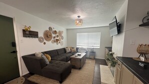 Ett modernt vardagsrum med en hörnsoffa, ett soffbord och dekorativ väggkonst. Det finns ett fönster med persienner, en tv som är monterad på väggen och en köksdel med en bänkskiva.