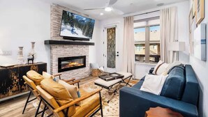 A modern living room with a fireplace, a large flat-screen TV, a ceiling fan, and a view of the outdoors through a window.