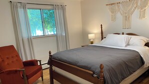 Une chambre avec un lit en bois, un fauteuil rouge, une fenêtre avec des rideaux et des décorations murales en macramé.