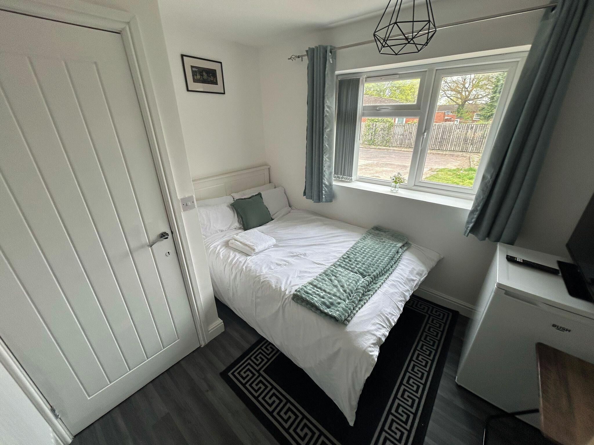 A bedroom with a bed, a window with curtains, a desk, and a radiator.