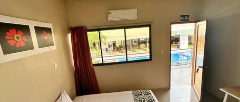 Habitación de hotel con cama, ventana con vista a una piscina al aire libre y una puerta con la leyenda 'SALIDA'.