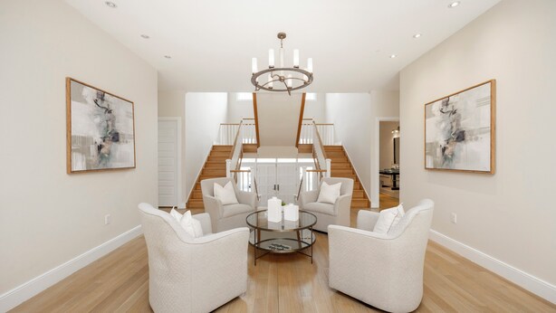 A spacious living room with a round glass coffee table, two white armchairs, a chandelier, and a staircase leading to another level.