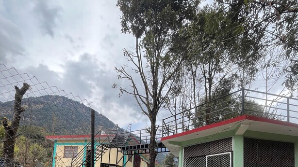 Un edificio con fachada verde y blanca, un espacio al aire libre cubierto con una valla metálica, y montañas al fondo.