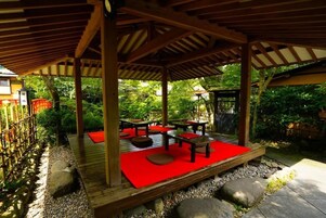 Un espacio de estar al aire libre con techo, mesas y bancos, rodeado por una terraza de madera y un jardín.