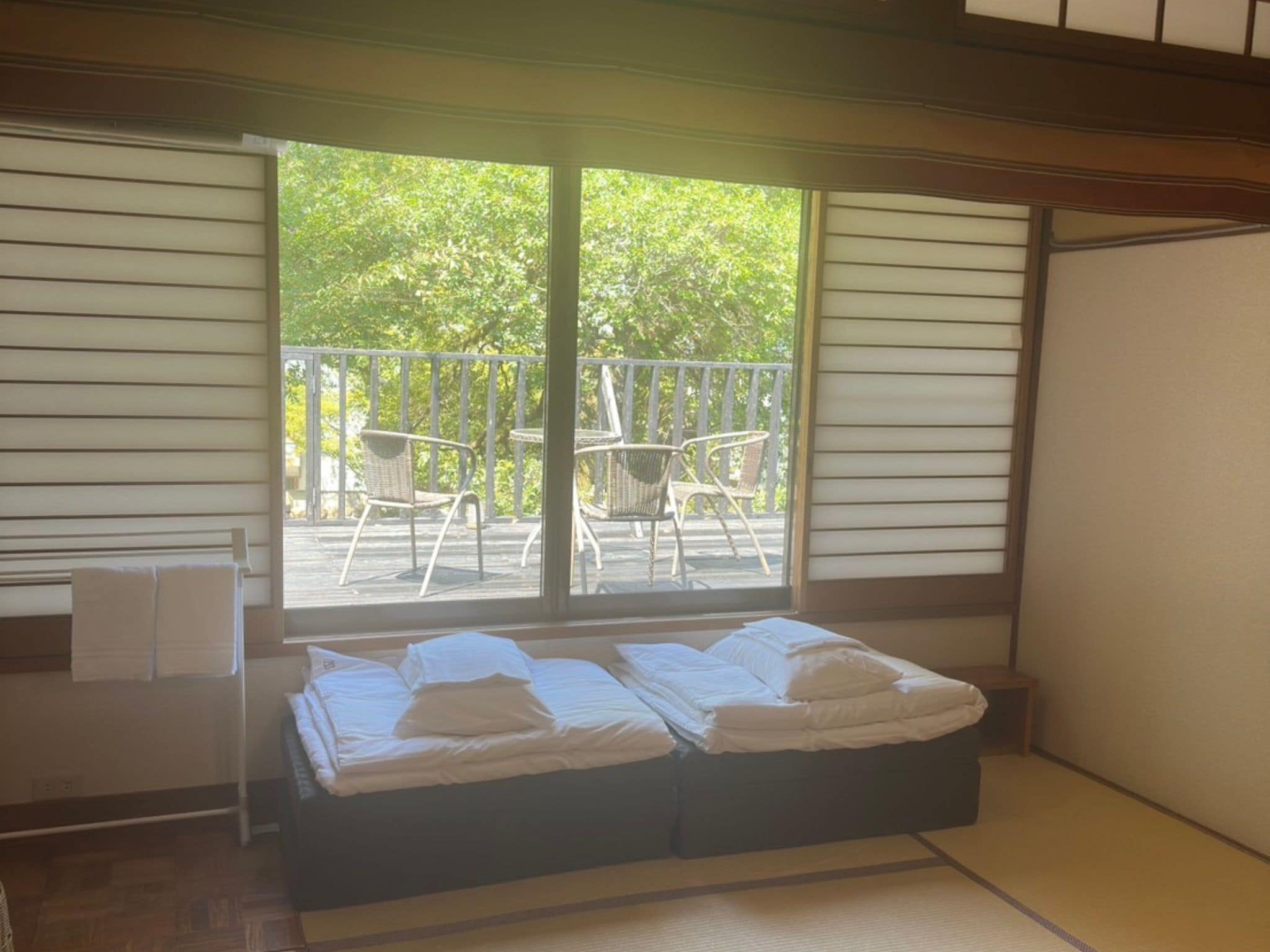 A room with a bed, a small table, and a chair. There is a balcony with a railing and a view of trees.