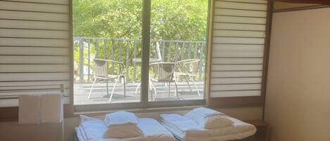 A room with a bed, a small table, and a chair. There is a balcony with a railing and a view of trees.