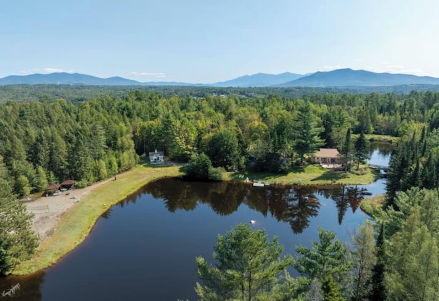 Une propriété au bord d’un lac, comprenant un chalet et un bâtiment plus grand, le tout entouré d’une forêt dense et avec des montagnes en toile de fond.