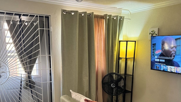 Una sala de estar con un sofá, un televisor montado en la pared, un ventilador de techo y una ventana con cortinas.