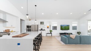 A modern kitchen with white cabinets, a central island with stools, a hanging light fixture, and a dining area with a blue sofa and a plant.