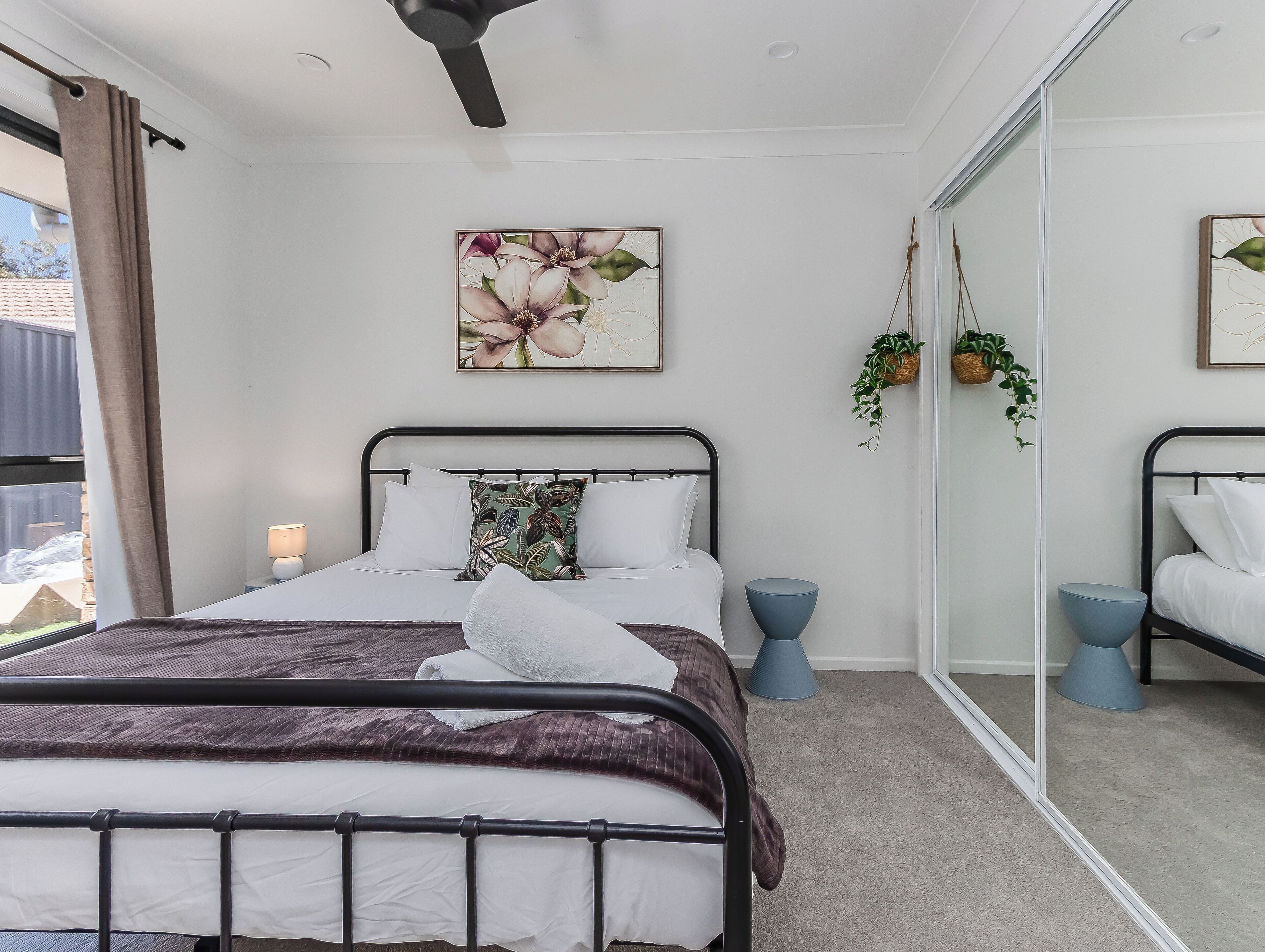 Une chambre à coucher comprenant un lit, des tables de chevet, un ventilateur de plafond, un grand miroir et une fenêtre avec des rideaux.