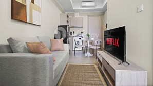 A modern living room with a grey sofa, a flat-screen TV, and a dining area in the background. - Serora by Kozystay - Kemayoran (Jakarta)
