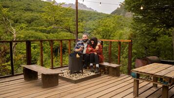 A couple sitting on a wooden deck with a fire pit, surrounded by lush greenery and mountains in the background.