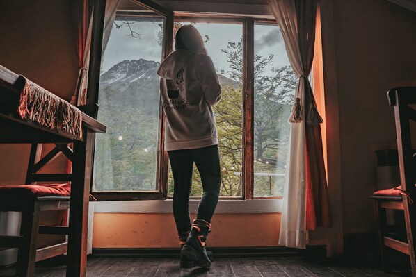 A person standing by a window with a mountain view, wooden furniture, and curtains.