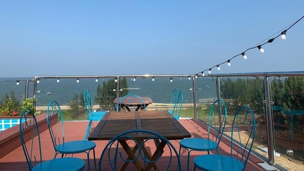 An outdoor dining area with a view of the sea, featuring a wooden table and blue chairs, and string lights overhead. - Aqua Resort Hai Tien - by BAYLUXURY (Hoang Tien)