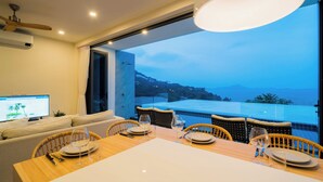 A dining area with a table set for four, a television on the left, and a view of the sea and mountains through the open door. - Staylar Seaview Pool Villa - Coral Cove, Koh Samui CC2 (Koh Samui, Coral Cove)