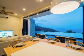 A dining area with a table set for four, a television on the left, and a view of the sea and mountains through the open door. - Staylar Seaview Pool Villa - Coral Cove, Koh Samui CC2 (Koh Samui, Coral Cove)
