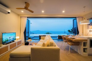 A modern living room with a sofa, TV, and dining area with a view of the sea.