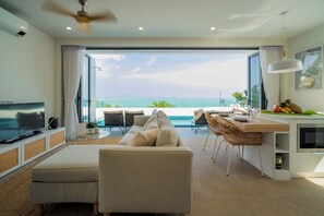 Una sala de estar moderna con un sofá, un comedor con mesa y sillas, una cocina con frutero, y un ventanal con vistas al océano.