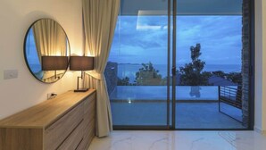 Habitació moderna amb un prestatge de fusta, un mirall rodó, dues làmpades i una gran porta corredissa de vidre que dona a un balcó amb vista al mar.