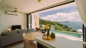 A modern living room with a large window, a ceiling fan, a sofa, a TV, and a dining table with fruit. - Staylar Seaview Pool Villa - Coral Cove, Koh Samui CC1 (Koh Samui, Coral Cove)