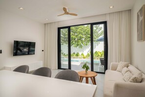 A modern living room with a dining area, a TV mounted on the wall, a ceiling fan, and a large sliding glass door leading to an outdoor area with a pool and greenery.