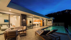 Une maison moderne dotée d’une piscine, de portes vitrées donnant sur un espace de vie comprenant un canapé et une table à manger, et d’une cuisine visible à travers les portes vitrées.