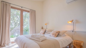 A bedroom with a large bed, two bedside lamps, a wooden nightstand, and a view of trees through the window.