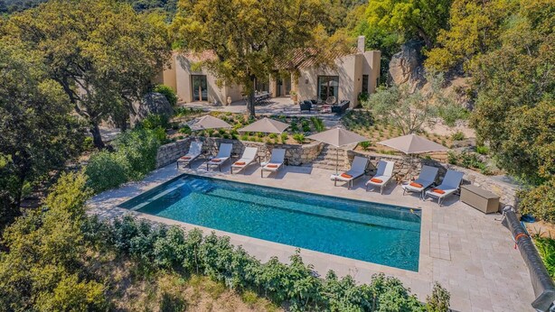 A pool with sun loungers and umbrellas, surrounded by greenery and a hillside.