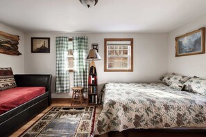 A bedroom with a red bed, a floral bedspread, a wooden floor, a bookshelf, and a window with checkered curtains.