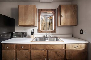 A compact kitchen with wooden cabinets, a stainless steel sink, and a window with a view of trees. - Lakefront w Water Toys, Deck, Fire Pit & Grill (Springfield)