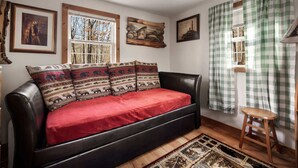 Une pièce chaleureuse avec un canapé en cuir noir, un plaid rouge et un tapis à motifs. Les murs sont ornés de tableaux encadrés, il y a une fenêtre donnant sur des arbres et un rideau à carreaux.