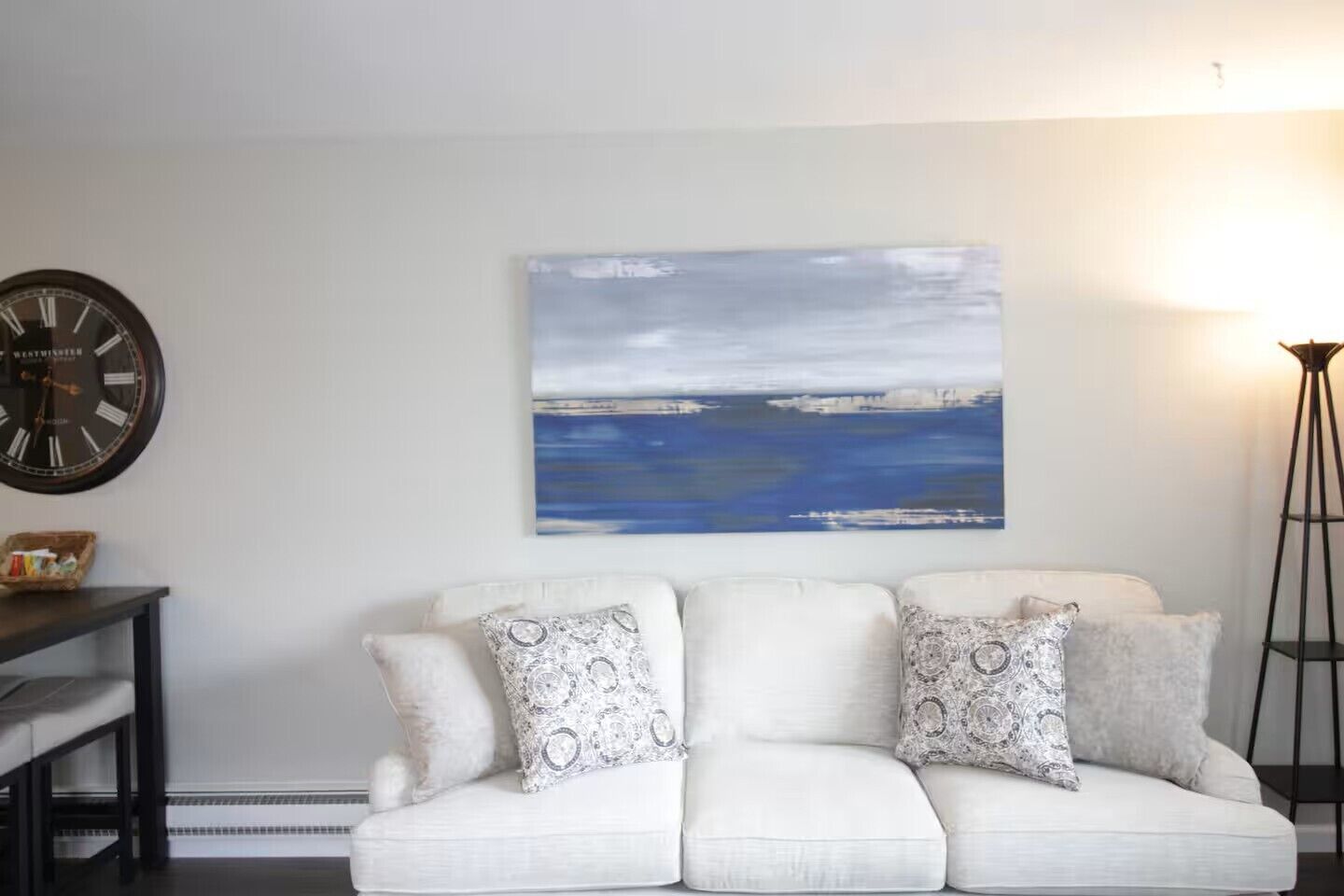 A living room with a white sofa, patterned cushions, a clock, and a painting of a blue seascape.