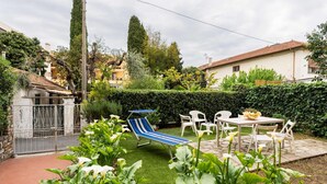 A patio with a table and chairs, surrounded by lush greenery and a small lawn.