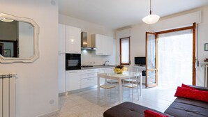 A modern kitchen with white cabinets, a dining area with a white table and chairs, a sofa with red cushions, and a TV in the corner.