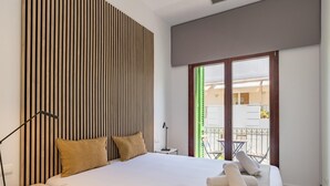 A modern bedroom with a large bed, a wooden slat headboard, a small bedside table, a floor lamp, and a view of a balcony with outdoor furniture.