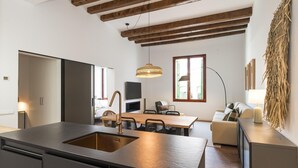 A modern kitchen with a central island, wooden beams on the ceiling, a dining area with a table and chairs, and a living space with a sofa and lamp.
