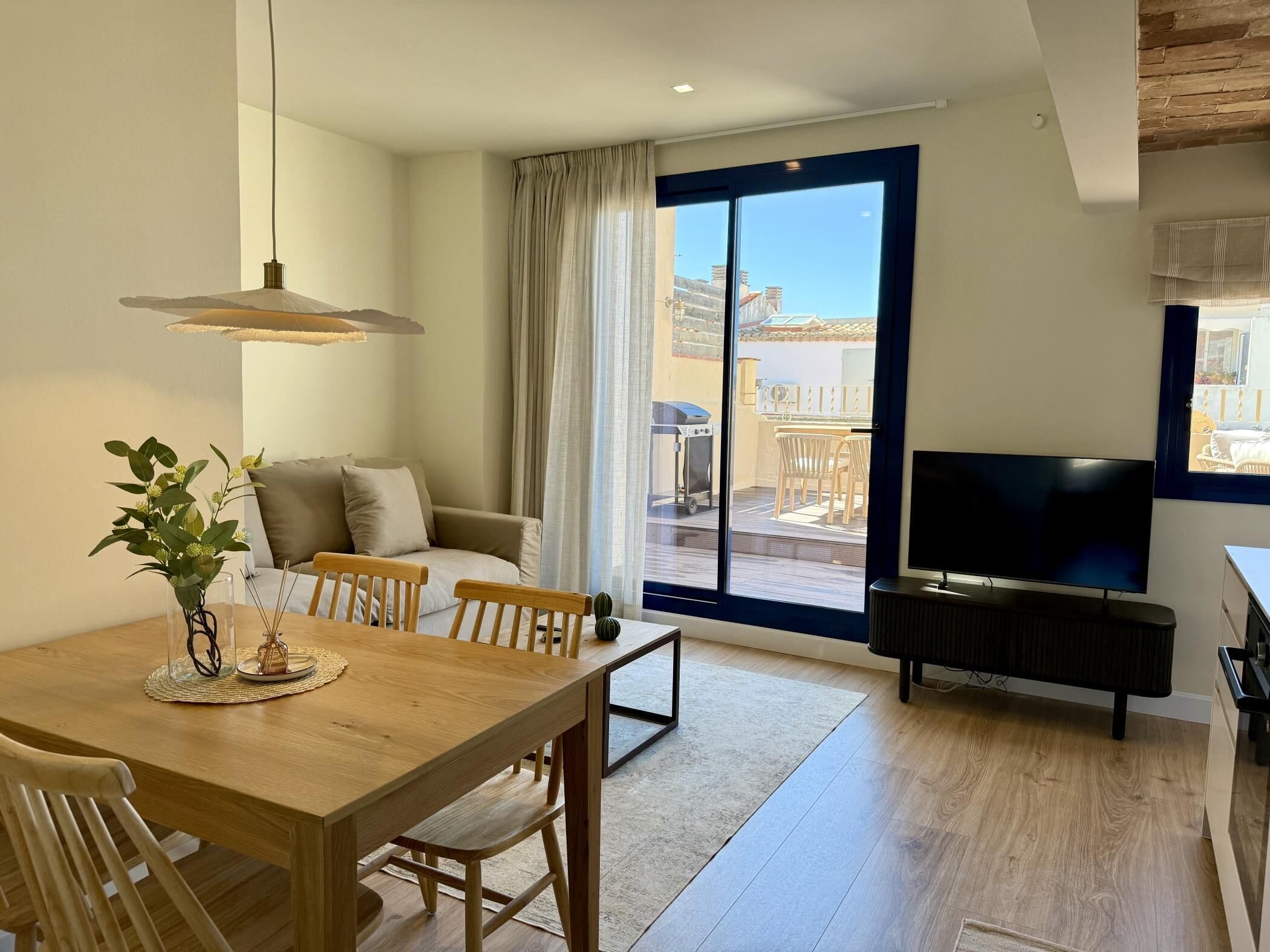 A modern living room with a wooden dining table, a sofa, a TV, and a balcony with a view.