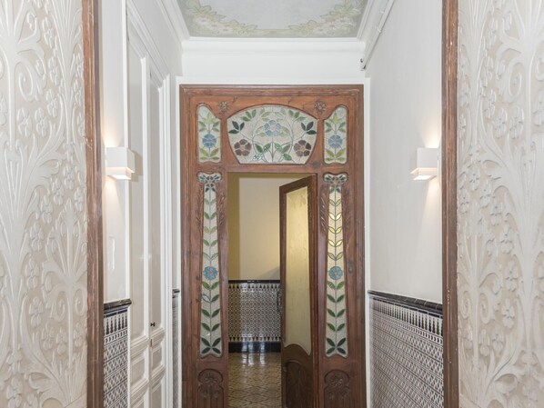 Un pasillo con puertas de madera decoradas, paredes con diseños y suelo de baldosas.