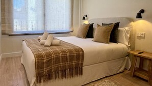 Un dormitorio con cama, almohadas, una colcha, mesita de noche, lámpara y ventana con cortinas.