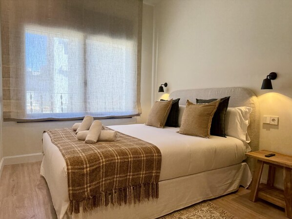 Ein Schlafzimmer mit Bett, Kissen, Decke, Nachttisch, Lampe und einem Fenster mit Vorhängen.