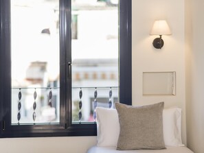 Un dormitorio con cama, una ventana con vistas a edificios, una lámpara de pared y un pequeño marco de fotos.